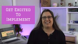 Image of a middle aged woman with brown hair in a black shirt sitting at a desk. The words "Get Excited To Implement" are written on the screen in white letters on a pink background.