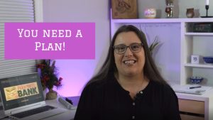 Image of a middle aged woman with brown hair in a black shirt sitting at a desk. The words "You Need A Plan!" are written on the screen in white letters on a pink background.